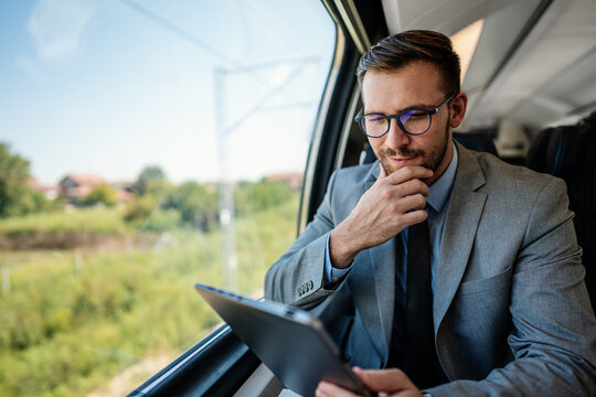 Professional male passenger in business suit analyzing digital data on touchscreen device while sitting by window on modern railway transport vehicle during morning trip.