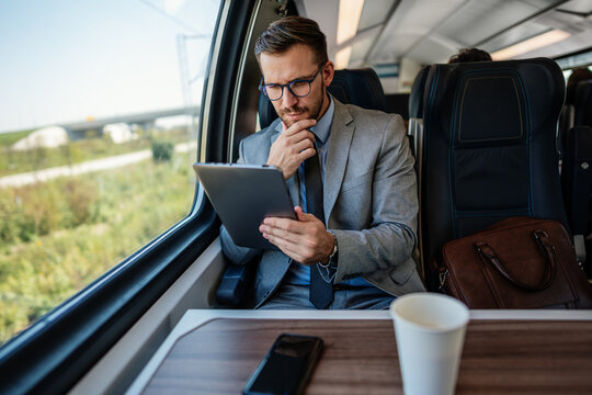Professional male passenger in business suit analyzing digital data on touchscreen device while sitting by window on modern railway transport vehicle during morning trip.