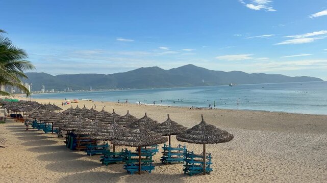 Panoramic view of Da Nang, Vietnam beach with rows of thatched umbrellas, sun loungers, swimmers, and mountains along the coastline