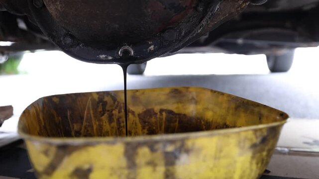 Close-up shot of dark, used differential oil draining from a car's rear differential housing directly into a yellow plastic catch basin during vehicle maintenance.