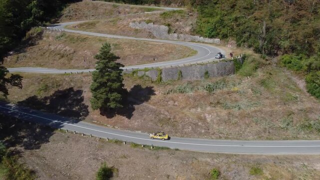 High aerial perspective of a yellow classic car navigating steep hairpin turns along a winding mountain road leading up from Bratca village in Romania, surrounded by lush forested hillsides.