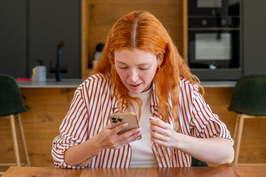Redheaded woman at a bright kitchen table reacts with surprise while reading a message on her smartphone, a candid moment of joy and connection in everyday modern home life.