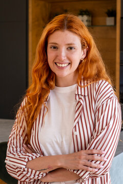 Confident redhead woman smiling in a casual striped shirt, portrait of a friendly young adult with crossed arms in a modern interior conveying warm, relaxed professionalism