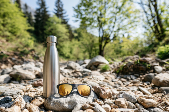 UV protective sunglasses with mirrored lenses placed beside a stainless steel water bottle on a rocky hiking trail, fresh outdoor atmosphere, natural daylight, commercial advertising aesthetic,,