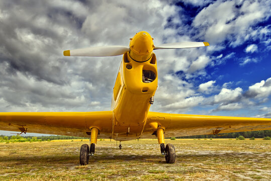 Yellow crop duster aircraft with propeller, parked on a dry grassy field, featuring a dynamic cloudy sky overhead