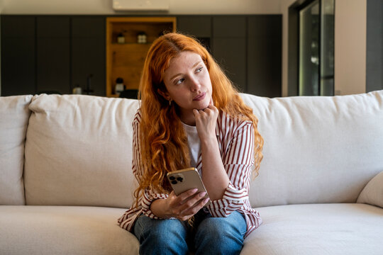 Young woman with red hair sits on a couch holding a smartphone, looking thoughtful and pensive while considering messages or plans in a bright modern living room, home moment.