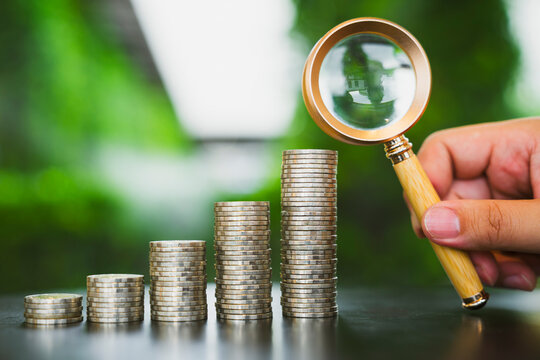 Financial audit and investment inspection concept with hand holding wooden magnifying glass examining rising coin stacks on table against green bokeh for accounting scrutiny and tax analysis.