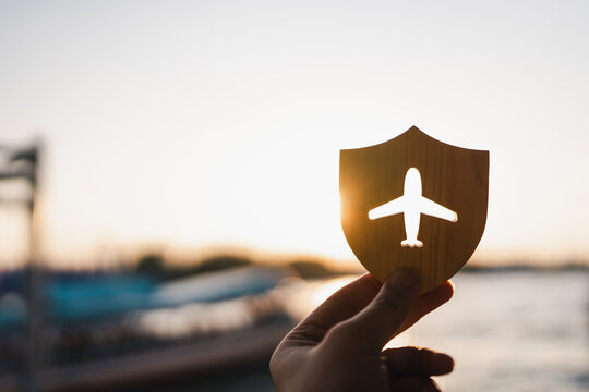 Travel insurance and evening flight protection concept with hand holding wooden shield with airplane cutout against golden sunset on water for holiday journey safety and tourist trip coverage.