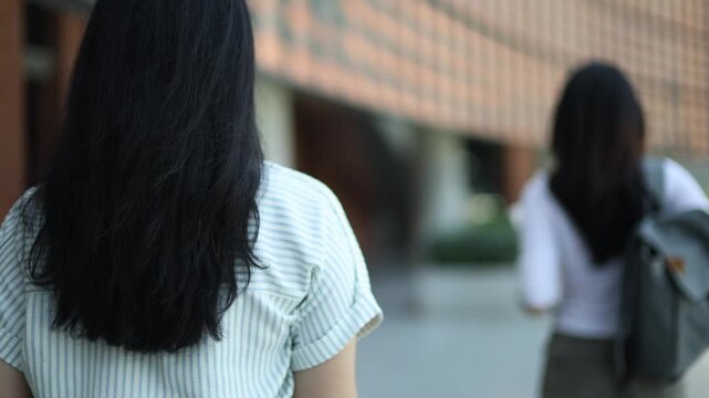 Asian female student carrying backpack and textbooks stands in front university building looking and smiling at the camera while fellow student walks away towards the building entrance.