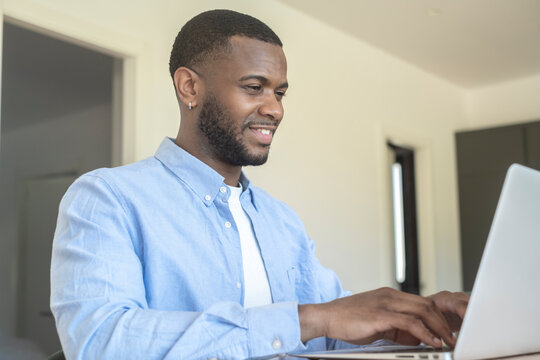 Young man working remotely on a laptop at home, focused and smiling while typing in a bright modern room, representing remote work, productivity, and casual professional lifestyle.