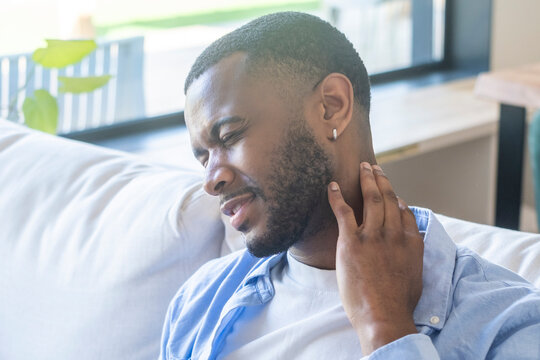 A young man on a sofa winces and rubs his neck, experiencing pain and stiffness at home by a sunlit window, showing discomfort, fatigue and the need for rest and self-care.