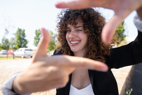 White woman with curly hair winking and framing hands outdoors