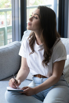 Young woman on a sofa by a window, looking thoughtful while writing notes in a notebook; natural light, casual outfit, journaling and a quiet reflective moment at home, peacefully.