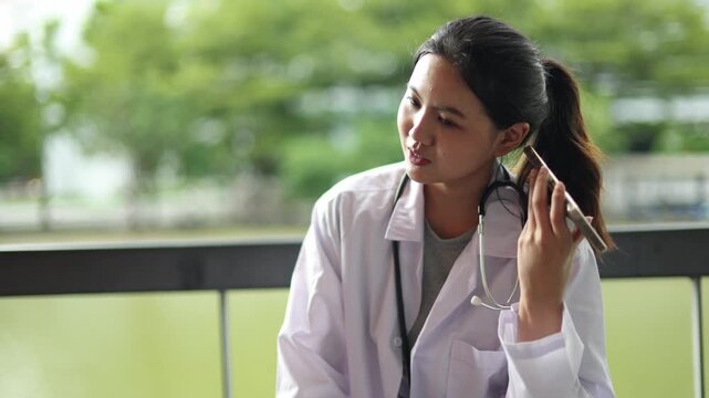 Asian female medical student dressed in her medical uniform sits in an academic building while chatting smartphone classmate who is waiting inside the modern facility.