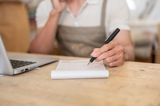 Close up of a man in apron hand writing on a receipt notebook at a wooden desk beside a laptop