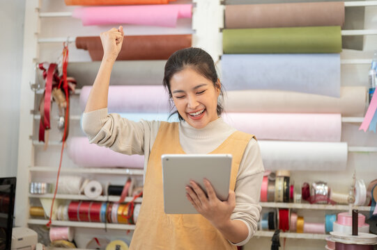 asian woman owner cheering success holding tablet in ribbon wrapping paper shop
