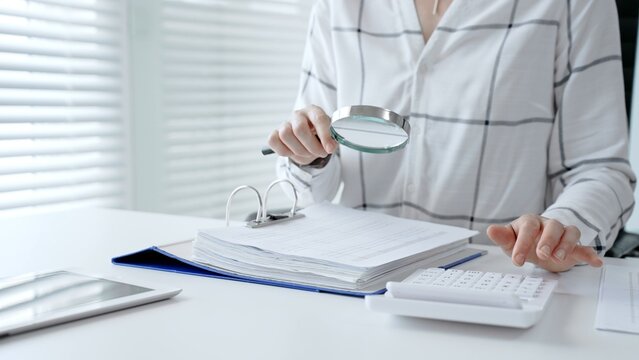 Accountant or auditor scrutinizing financial legal documents with a magnifying glass while using a calculator, performing business tax analysis and detailed investigation