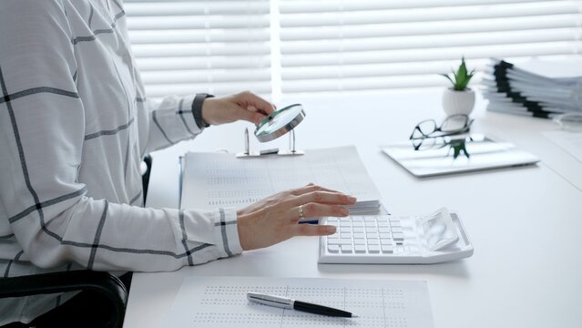 Financial professional carefully reviewing business documents with a magnifying glass while calculating numerical data, demonstrating diligent tax preparation and detailed audit analysis