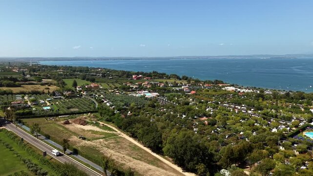 Lazise at Lake Garda in Italy with vineyards, olive groves, traffic on lakeside road, terracotta roof houses, campsites among trees, blue lake stretching to hazy shore. Aerial drone rising wide shot