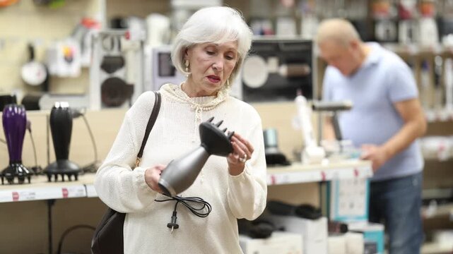Elderly woman stands in a store with household appliances and carefully chooses a hairdryer with different styling attachments . High quality 4k footage