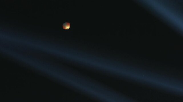 paper balloon glows small and orange against black sky, faint blue cloud bands crossing the frame behind it, alone at altitude