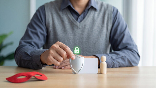 Identity theft protection concept, man placing silver shield with green padlock in front of blank card and wooden figure while red thief mask lies on desk for privacy and fraud prevention