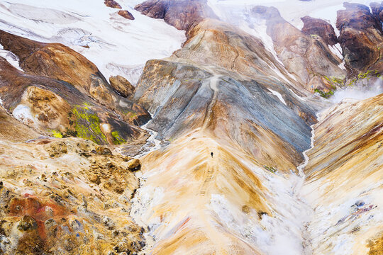 Aerial view of Kerlingarfjoll with colorful rhyolite mountains, geothermal steam, snow patches, and a hiker on a trail in Hrunamannahreppur, Iceland.