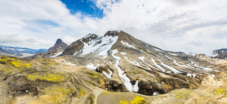 Aerial view of Kerlingarfjoll mountain range with snow-capped peaks, volcanic terrain, and vibrant yellow moss under a cloudy sky in Hrunamannahreppur, Iceland.