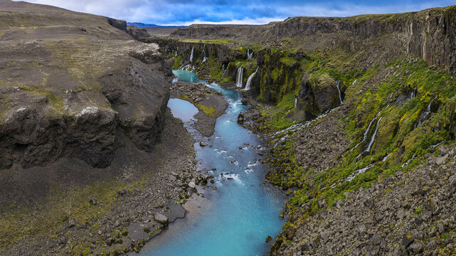 Aerial view of Sigoldugljufur canyon featuring a vibrant turquoise river flowing through deep volcanic cliffs adorned with cascading waterfalls and lush green moss in Iceland.