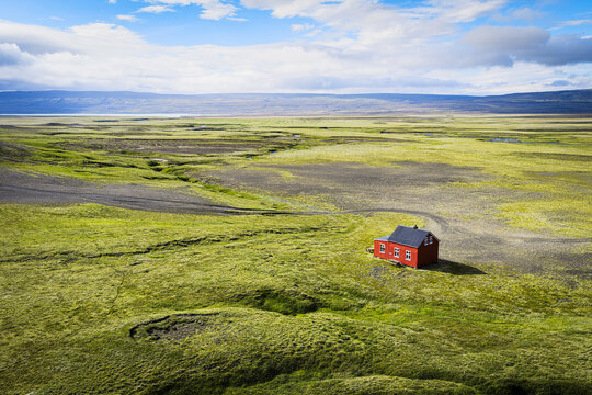 Aerial view of a solitary red house set within a vast, vibrant green landscape under a bright blue sky in Iceland.