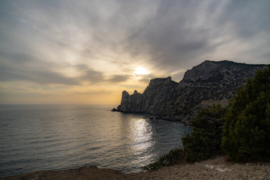 Dramatic coastal cliffs rise above dark ocean waters reflecting a sunset sun path under a cloudy sky