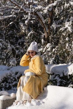 Young woman wearing yellow coat and white hat sitting on snow covered bench in park during sunny winter day