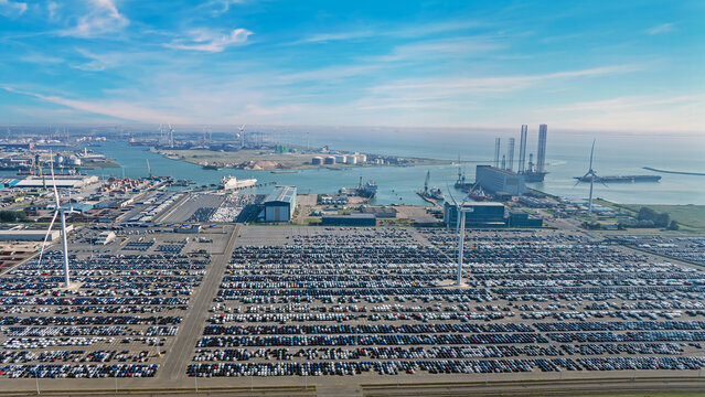 Aerial view of a vast car terminal at the port with thousands of parked vehicles, industrial infrastructure, wind turbines, and harbor facilities in Vlissingen, Zeeland, Netherlands.