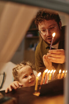 Vertical shot of Jewish Caucasian young adult father lighting Hanukkah menorah candles with female child daughter, suitable for holiday advertising campaigns and cultural family editorial