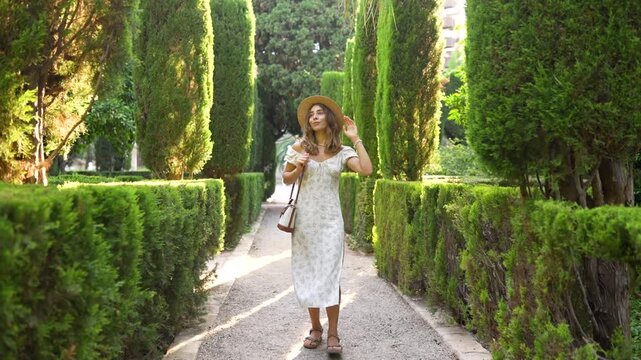 Woman in dress walks along path in beautiful garden, surrounded by greenery. Woman, wearing hat, enjoys an outdoor stroll. Concept beauty, leisure, or weekend getaway.