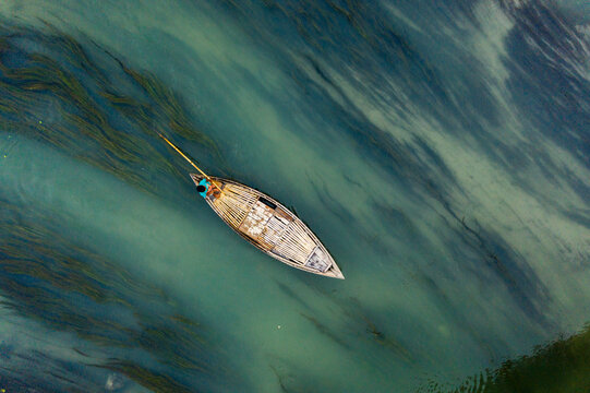 Aerial view of a traditional wooden boat with a fisherman using a long pole to navigate through clear river water with aquatic plants in Sonatola Upazila, Rajshahi Division, Bangladesh.