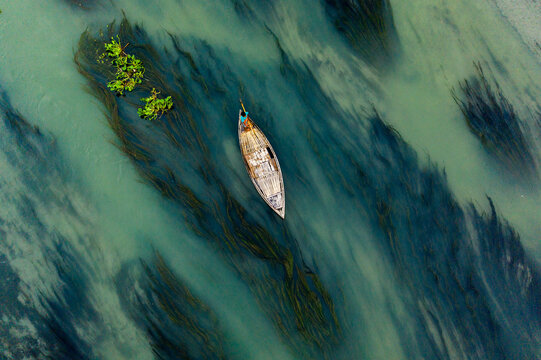 Aerial view of a small wooden boat navigating through clear river water filled with aquatic plants, Sonatola Upazila, Rajshahi Division, Bangladesh.