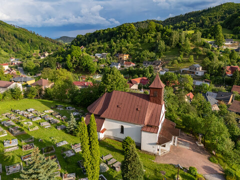 Aerial view of the Parish Church of Assumption of Virgin Mary in Riecka surrounded by a cemetery, lush green hills, and village houses in Riecka, Banska Bystrica Regio, Slovakia.