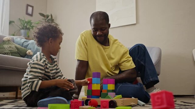 African American father and young son building colorful toy block tower on floor at home. Clip fits parenting campaigns, early childhood education and toy advertisements