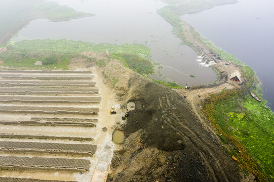 Aerial view of the brick kiln site featuring long rows of drying bricks, piles of soil, and water channels near the river in Aminbazar, Dhaka Division, Bangladesh.