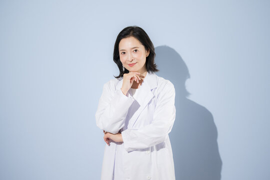 [水色背景]白衣を着て考えるポーズをする女性医師、笑顔でカメラ目線日本人ドクターのポートレート