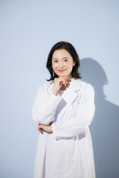 [水色背景]白衣を着て考えるポーズをする女性医師、笑顔でカメラ目線日本人ドクターのポートレート