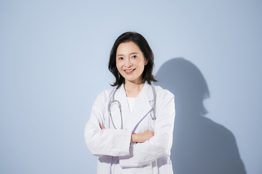 [水色背景]聴診器をつけて白衣を着て考える腕を組むポーズをする女性医師、笑顔でカメラ目線日本人ドクターのポートレート