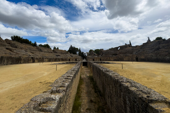 ancient roman amphitheater ruins in serene outdoor setting with stone walls and sand floor, wide angle perspective from within the structure, historical and travel context.