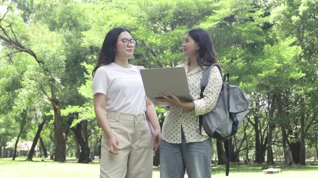 Two knowledgeable and self assured Asian female students stand on a university walkway amidst nature wearing backpacks carrying laptops review lessons together while conversing warm genuine smiles.