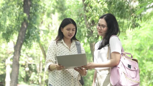 Two knowledgeable and self assured Asian female students stand on a university walkway amidst nature wearing backpacks carrying laptops review lessons together while conversing warm genuine smiles.