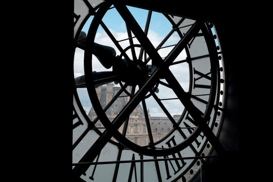 Large iconic clock face of mus&eacute;e d'orsay with roman numerals, framing historic architecture of louvre museum under a cloudy sky