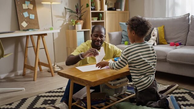 African American father and young son making, flying paper planes indoors during playful leisure. Clip works for parenting campaigns, fatherhood storytelling, and early childhood education