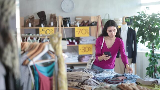 Young woman scans QR code and label of trousers or pants using mobile phone camera. Customer client is interested in country of origin of product, checks certificate on consumer protection website. 