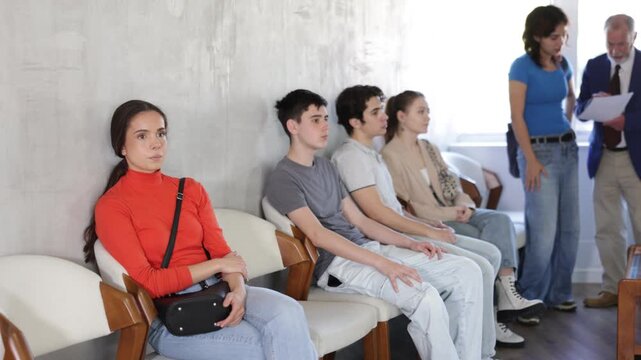 Relaxed young visitors sitting in a queue while senior manager speaking to one in background 